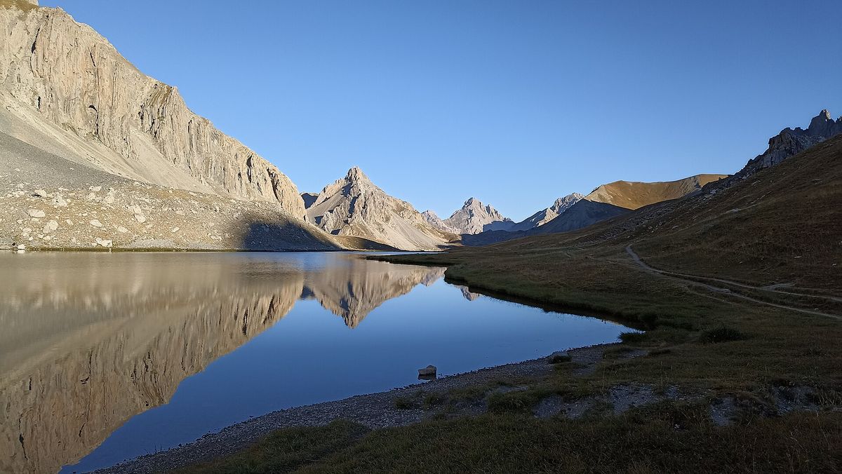 Le lac de l'Oronaye au petit matin, les falaises se reflétant dans l'eau calme, le sentier longeant la rive
