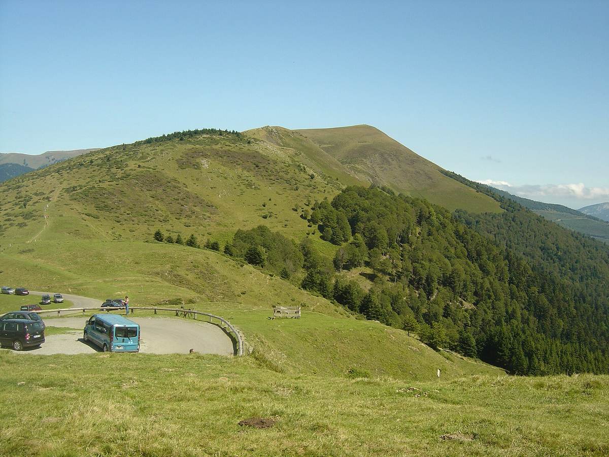 Le sommet herbeux et arrondi du Plo del Naou au-dessus d'un parking, avec une forêt de sapins sur son flanc